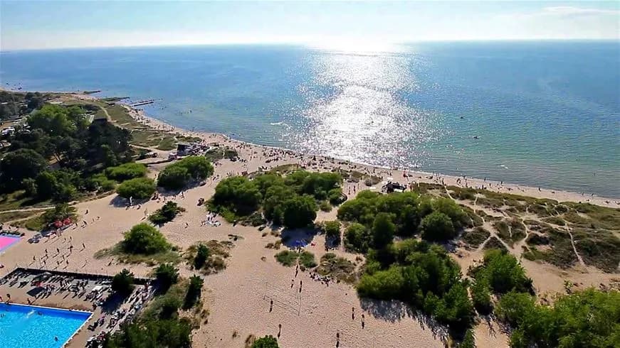 Flygbild över Tofta camping med stranden i förgrunden och ett stort antal människor som njuter av sommarvädret. Havet syns bakom stranden, med klarblått vatten och vågor som rullar in mot kusten.