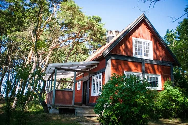 En lantlig stuga omgiven av gröna träd och växtlighet på Tofta Camping, med ett soligt vårlandskap som bakgrund.