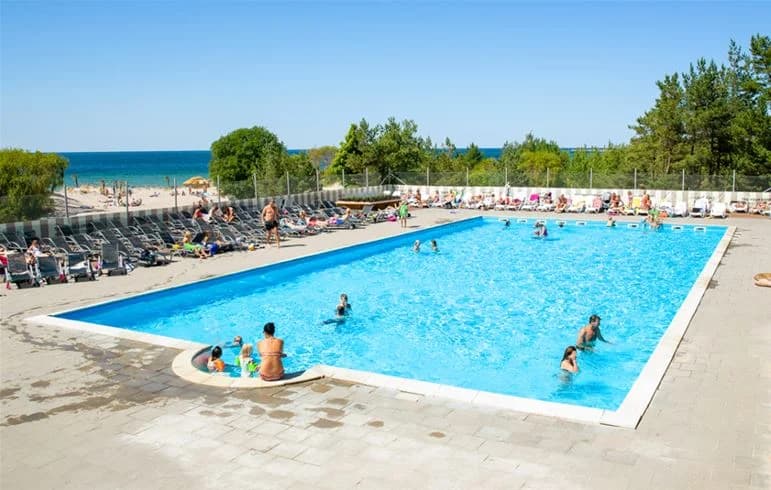 En solig dag vid Tofta camping, med besökare som njuter av semesteraktiviteter. Flera personer är samlade runt en utomhuspool, där några simmar och andra kopplar av på solstolar. I bakgrunden syns semesteranläggningens bekvämligheter och sandstranden ligger nära, vilket ger en känsla av sommar och rekreation.