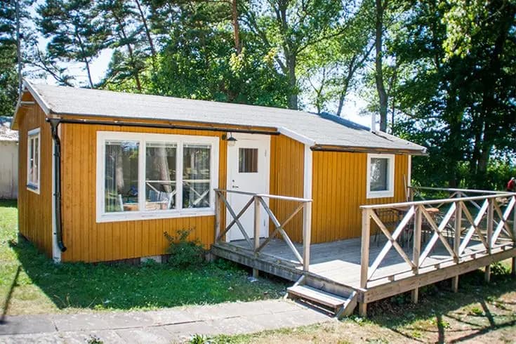 En trästuga på Tofta Camping omgiven av en grönskande trädgård med en veranda.