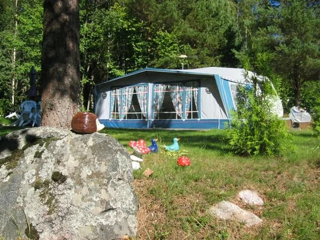 En tältplats i en skogsmiljö på Kapellskärs camping, med ett tält och en skyddande presenning som ger skugga. Campingstolar står utplacerade i det natursköna området, omgivet av träd och grönska.