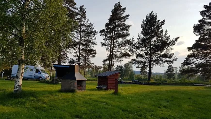 En grön campingplats med vidsträckt gräsyta och flera träd. En enkel byggnad eller skjulet är synligt i bakgrunden. Platsen ger en lantlig känsla och omges av öppna ytor och växtlighet.