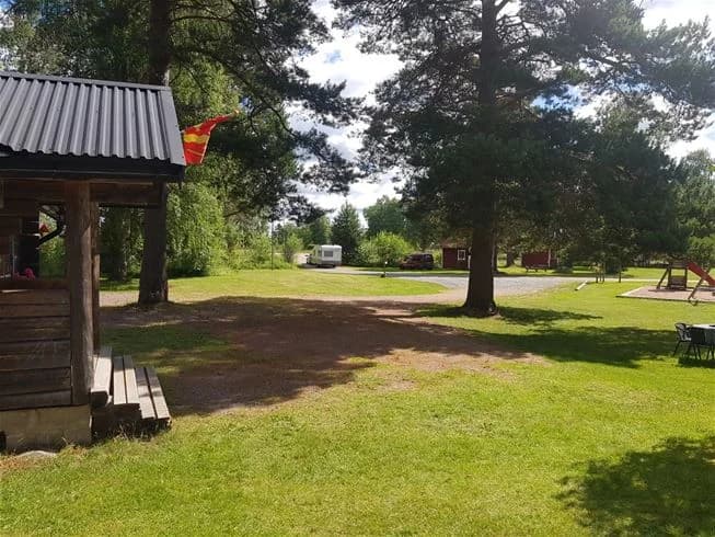 En grön campingplats med träd, gräs och stugor samt skuggiga ytor.