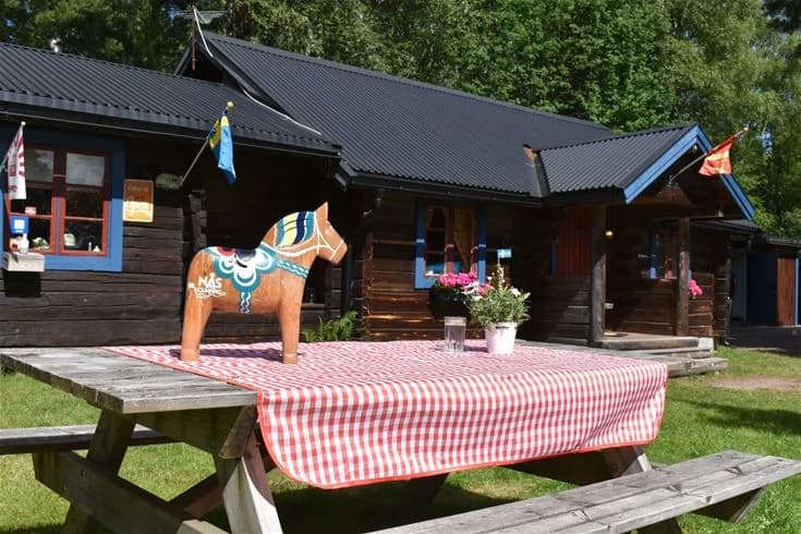 Bild av en timmerstuga omgiven av skuggiga träd i en trädgårdsmiljö. En utomhusplats med bord och stolar erbjuder en plats för rekreation och avkoppling.