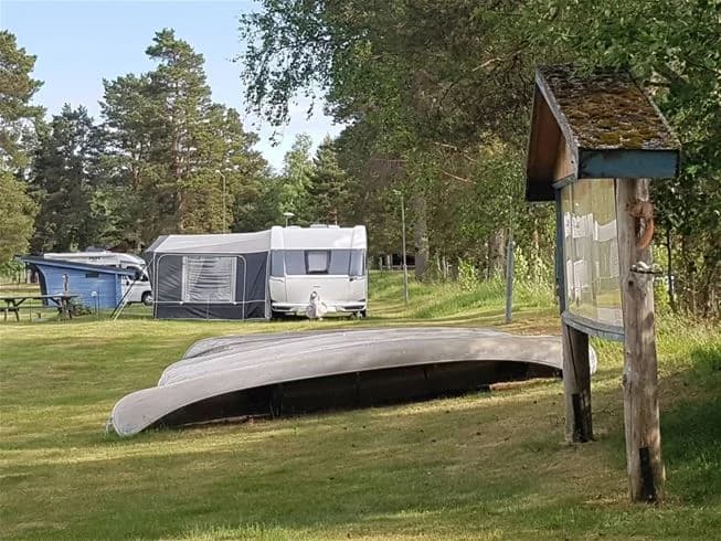 En grön campingplats med gräs och skuggande träd, där tält står uppställda på en öppen gräsmatta. I bakgrunden finns några trädgårdsbyggnader.