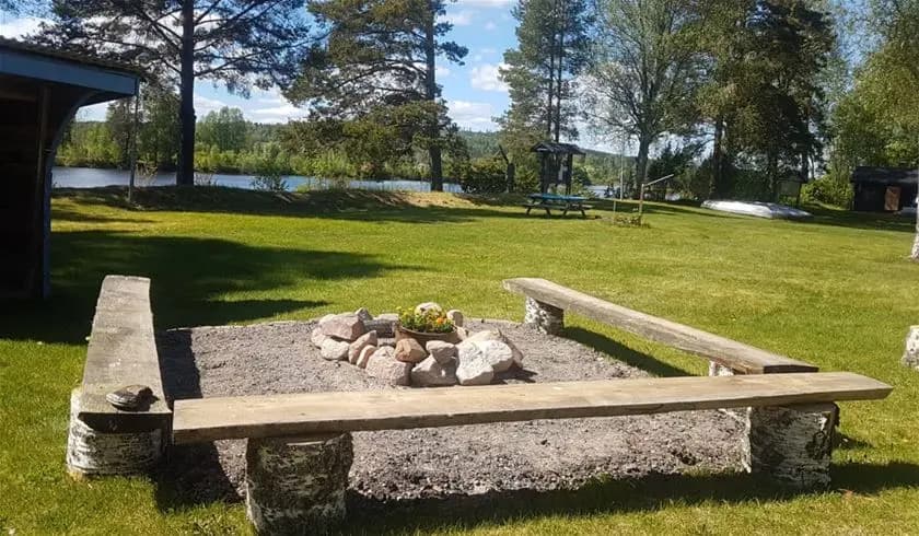 En träpicknickbänk står i skuggan omgiven av grönska vid Nås camping, med parkmiljö i bakgrunden.