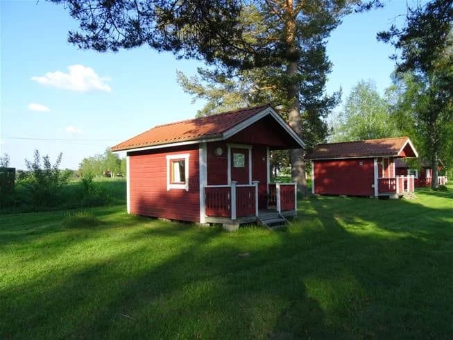 En grön äng med flera små byggnader, inklusive stugor och skjul, omgiven av välskött gräsmatta och trädgård i ett campingområde.