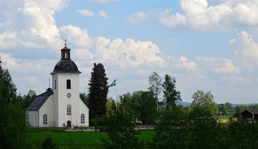 En liten kapell ligger vid kanten av en stilla damm, omgiven av tät skog med vintergröna träd i ett våtmarksområde vid Nåås camping.