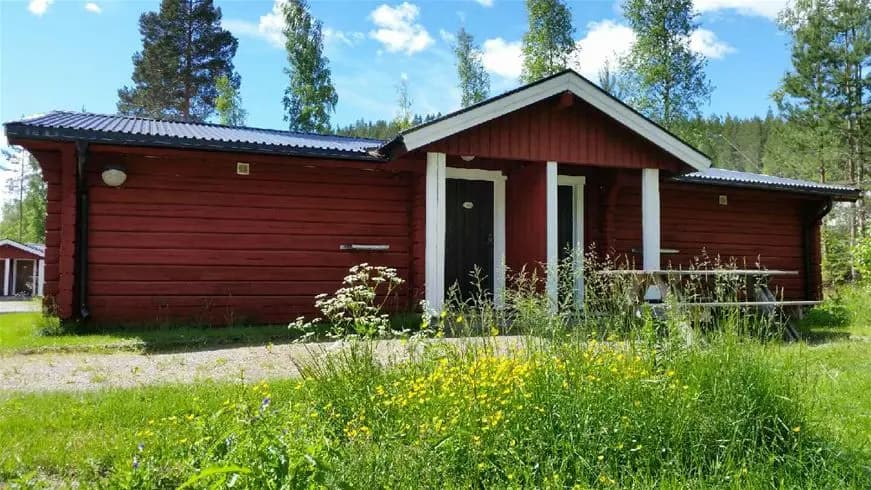En mysig stuga med rött tak, omgiven av grönskande landskap och små trädgårdsbyggnader, belägen i det natursköna Bjursås Berg & Sjö.