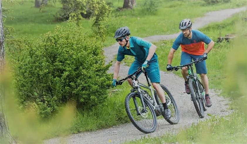 En cyklist med cykelhjälm cyklar på en stig i en naturskön miljö, omgiven av grönska och träd. Cykeln är fullt utrustad med alla nödvändiga delar såsom hjul och styre. Besöksmålet är Bjursås Berg & Sjö.