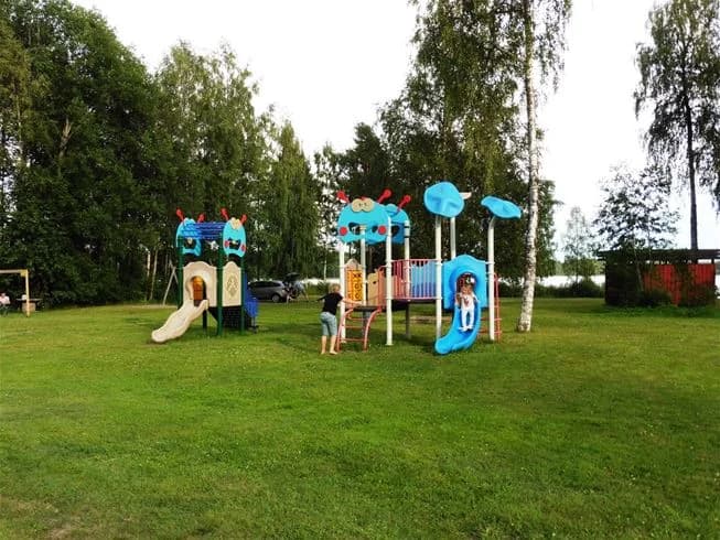 En grupp människor njuter av fritiden i en park vid Bjursås Berg & Sjö. Området visar lekutrustning och en grön gräsmatta med skuggiga platser för avkoppling. Perfekt för lek och semesteraktiviteter.