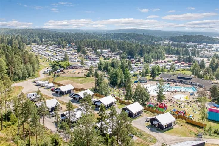 En flygbild över Skellefteå Camping, med träd, en pool och stugor som sprider ut sig i det natursköna landskapet, omgiven av grönska och ett rekreationsområde.