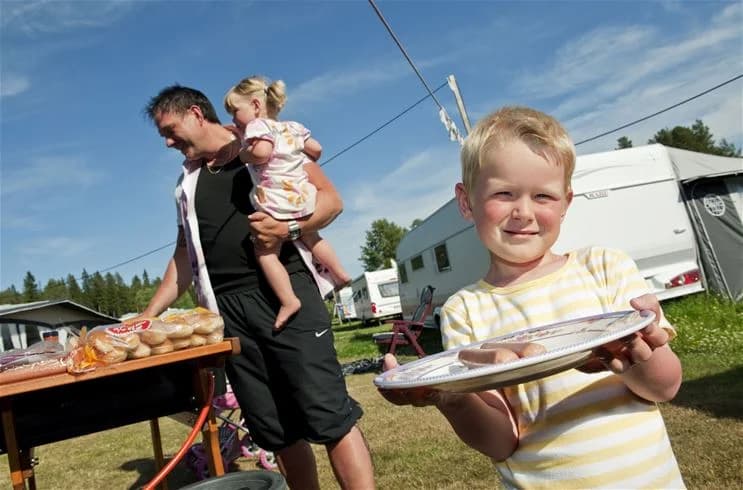 En familj äter lunch utomhus vid Skellefteå camping. Barnen ser glada ut när de njuter av sin måltid vid ett dukat bord med tallrikar och bestick. I bakgrunden syns campingens natursköna miljö.