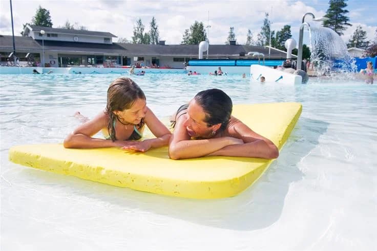 Besökare njuter av solen vid Skellefteå camping, där flera personer badar och leker i en stor utomhuspool. Det är en livlig atmosfär, med semesterfirare som kopplar av och har roligt i sina badkläder, omgivna av gröna områden och semesteranläggningar.