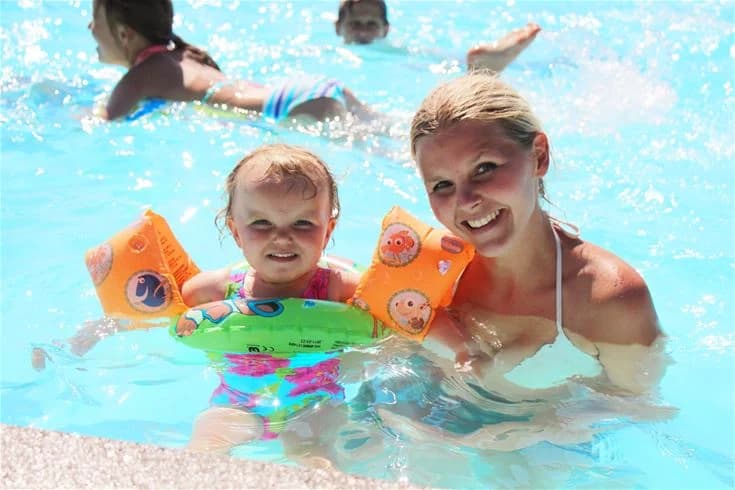 En grupp glada människor i badkläder njuter av en dag vid en pool på Skellefteå camping. De ler och umgås omgivna av uppblåsbara badleksaker och säkerhetsutrustning, vilket skapar en avslappnad och rolig atmosfär.