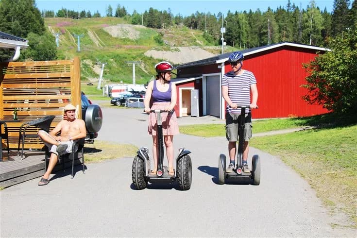 En grupp människor åker på Segways vid Skellefteå camping. De ser glada ut och njuter av sin fritid i soligt väder, några av dem bär solglasögon. En perfekt dag för semester och rekreation.