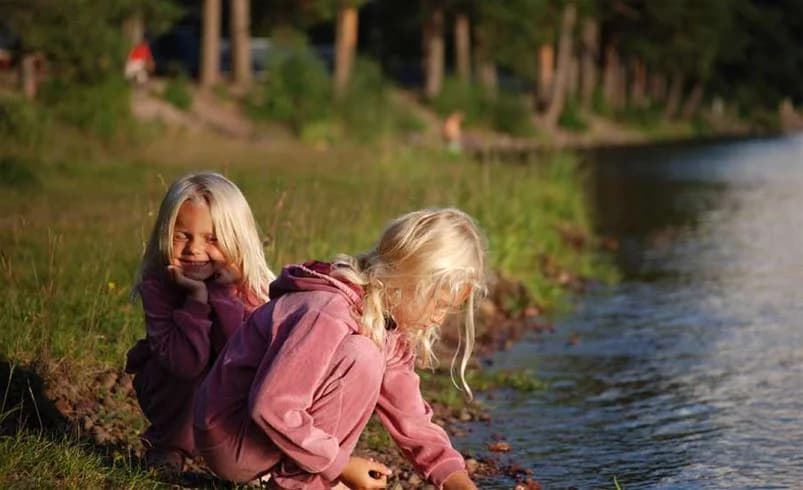 En bild av Älvdalens camping med en vacker sjö i förgrunden, där träden reflekteras i vattnet. Människor njuter av naturen, inklusive ett barn som leker nära strandkanten. Omgivningen domineras av skog och våtmarker, vilket ger en känsla av lugn och rekreation.