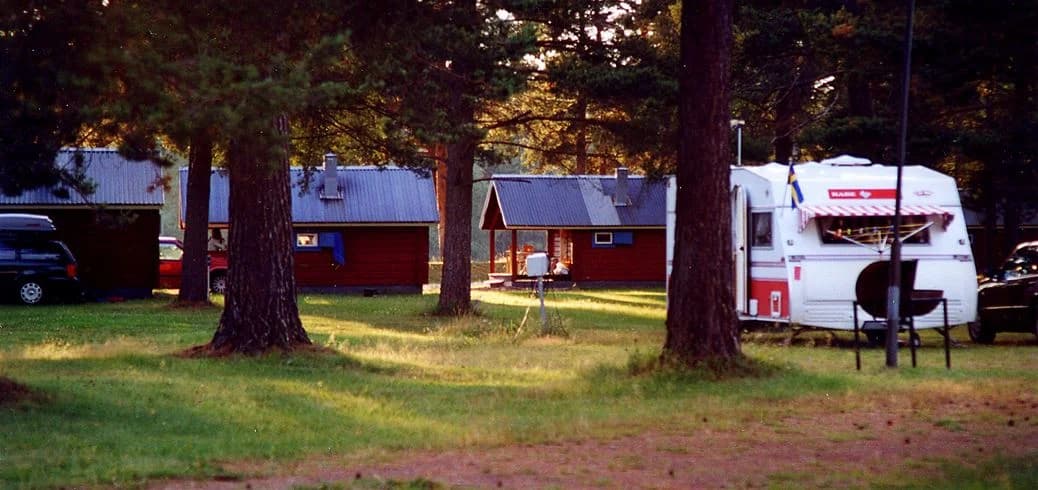 En grön campingplats omgiven av träd med en husvagn och en timmerstuga på en gräsmatta.