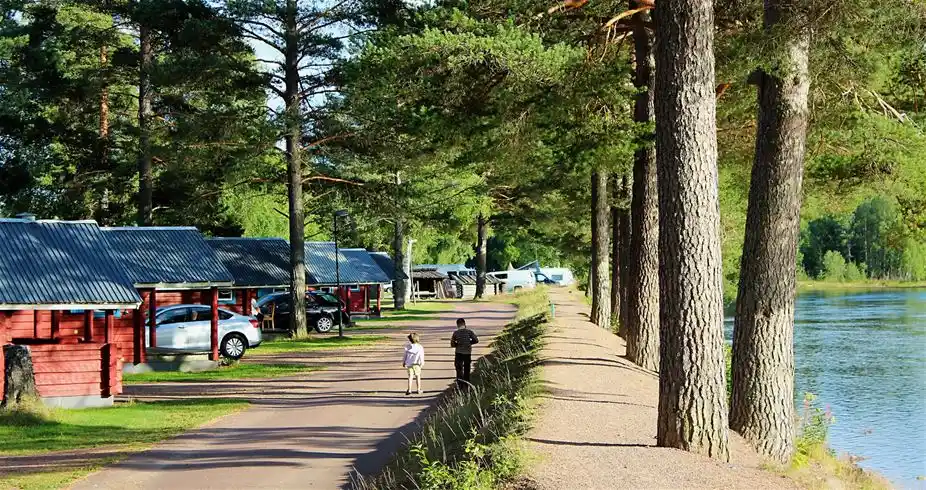 En timmerstuga omgiven av skuggiga träd vid en liten sjö på Älvdalens camping, med utemöbler placerade i det gröna gräset.