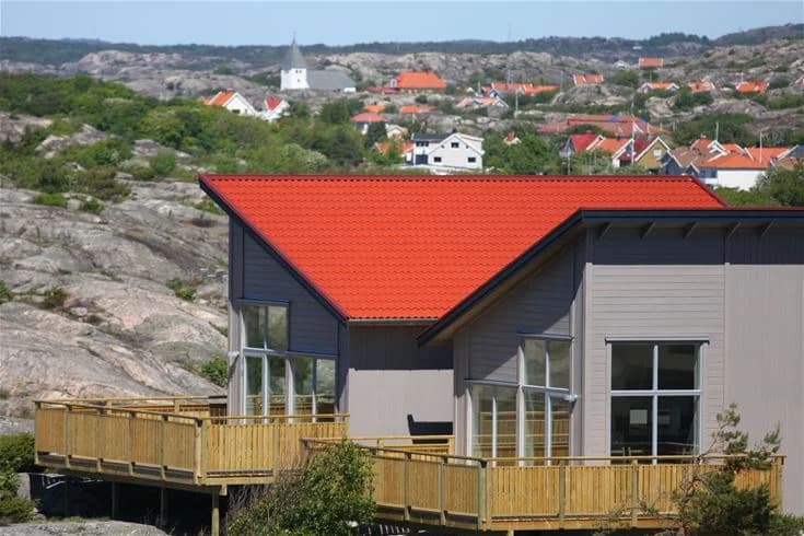 En mysig stuga vid Hav & Logi Skärhamn, med träfasad och stort däck med sittplatser, omgiven av naturskön miljö i dagsljus.