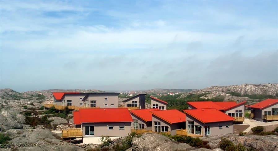 En pittoresk stuga med rött tak och vit fasad omgiven av karga klippor och grönska, belägen i det natursköna landskapet vid Hav & Logi Skärhamn.