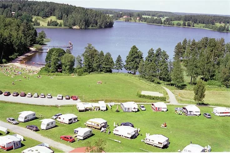 Bild på Malmköpings Bad & Camping: En lugn rekreationsplats vid en sjö omgiven av grönskande gräsmattor och ängar. Perfekt för fritidsaktiviteter och avkoppling i en naturskön miljö.