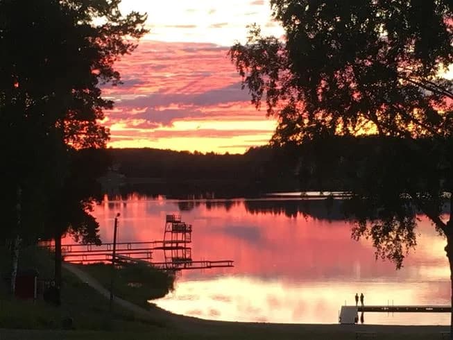 En somrig kvällsvy vid Malmköpings bad & camping med solnedgångens varma orangea sken som reflekteras i sjöns stilla vatten.