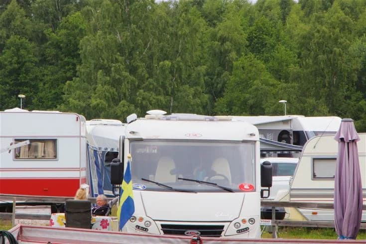 Bild av en stor husbil parkerad vid en campingplats i Malmköping, med omgivande skog och klarblå himmel i bakgrunden.