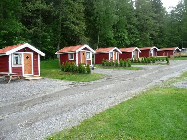 En mysig stuga bland grönskande träd och buskar, belägen vid Malmköpings Bad & Camping. I förgrunden syns en grusad uppfart som leder till stugan och omgivningarna präglas av en fridfull parkmiljö med inslag av barrträd och välskötta gräsytor.