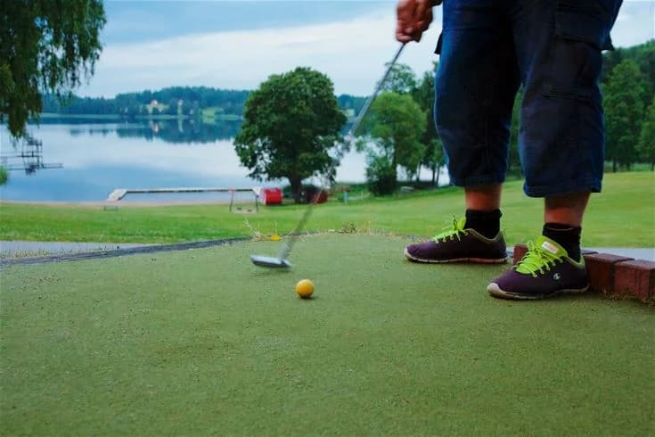 En grönskande campingplats i Malmköping med gräsytor där människor njuter av fritidsaktiviteter, inklusive minigolf. Besökare syns i avslappnade kläder och sandaler, omgivna av leksaker och golfklubbor på den välskötta gräsmattan.