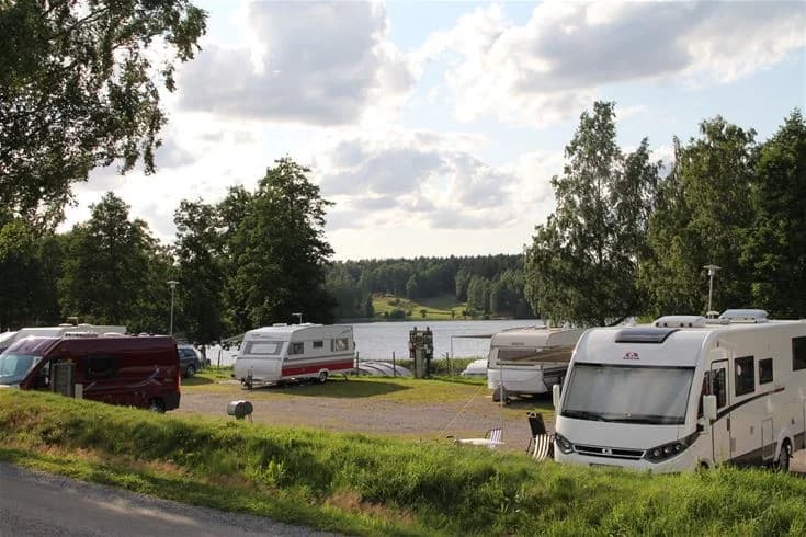 En campingplats med husbilar och husvagnar parkerade bland träd, med en sjö i bakgrunden och flera fordon synliga på området.