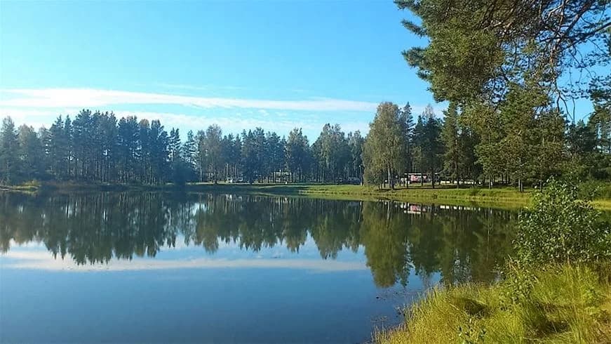 Vy över Vivstavarvstjärns camping med himmel och blått vatten i bakgrunden, omgiven av gröna träd och naturskön miljö.