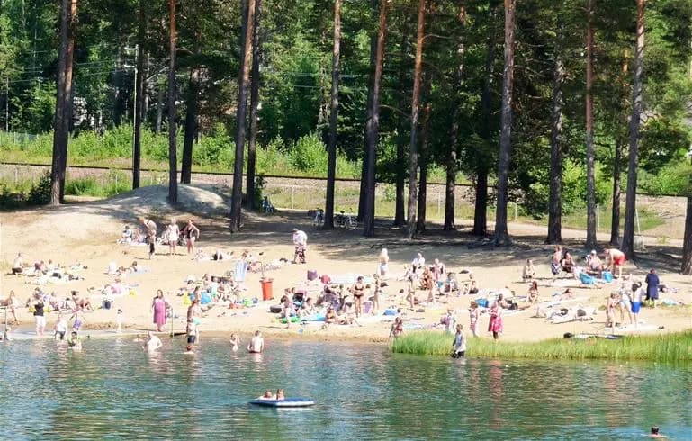 En sjö med en sandstrand vid Vivstavarvstjärns camping. Personer njuter av fritidsaktiviteter och avkoppling vid vattenkanten en solig dag.