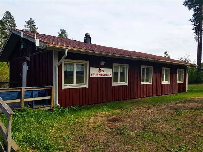 Bild på en charmig stuga vid Vivstavarvstjärns camping. Stugan har en träfasad med en snedtak och en trädörr, omgiven av en välskött gård och grönska.