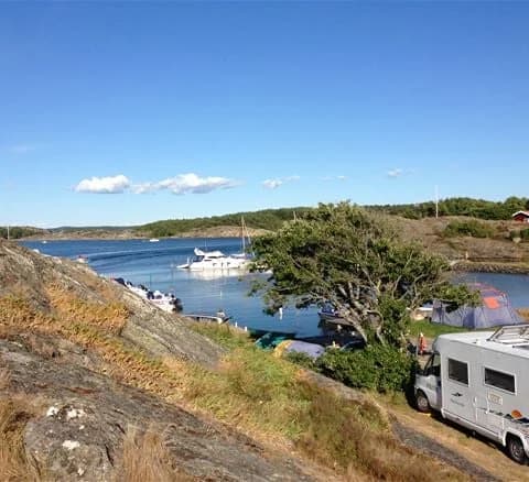 En idyllisk vy över Malö Camping med en kustlinje där småbåtar ligger förtöjda vid en brygga. Bakom båtarna syns en lugn vik med grönskande öar i fjärran under en klarblå himmel.