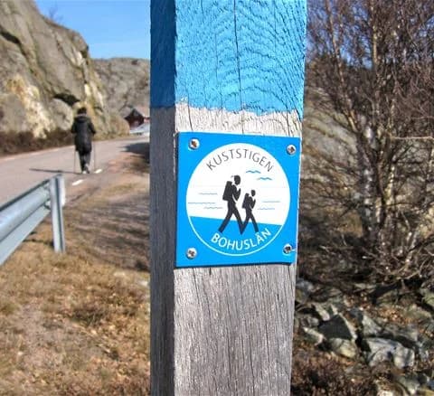 En välkomnande skylt vid Malö Camping omgiven av natur, en person med ryggsäck syns i bakgrunden, vilket antyder utomhusäventyr och rekreation.