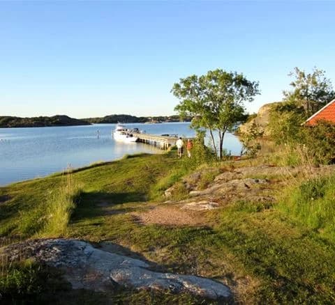 Vy av Malö Camping med en vacker kustlinje, där en båt syns på det stilla vattnet. Stranden sträcker sig längs sjökanten, omgiven av frodig grönska och en klar himmel i bakgrunden.