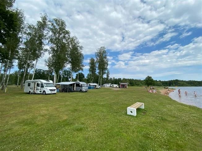 En solig dag vid Hultsfred Strandcamping med en klarblå himmel och vita cumulusmoln. Kustlinjen sträcker sig längs en lugn sjö med en sandstrand där besökare kan njuta av friluftsliv och rekreation.