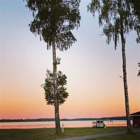 En solnedgång över ett naturskönt landskap vid Hultsfred Strandcamping, med en vid horisont och himmel i varma skymningsfärger. Träden skapar skuggor på marken och ger en stämningsfull känsla av kväll.