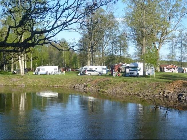 Vy över en lugn vattenmassa omgiven av frodig grönska och en gräsklädd strandkant, med inslag av kanaler och småvatten i bakgrunden vid Hultsfred strandcamping.