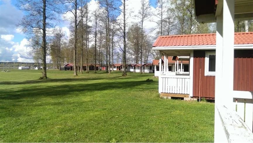 Bild av Hultsfred strandcamping: En grön gräsmatta omgiven av ett öppet landskap med en liten stuga i bakgrunden, vilket ger en lantlig och avkopplande känsla.