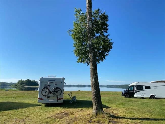 En campingplats vid Hultsfred strandcamping med flera uppställda husbilar och husvagnar. Parkerade fordon, inklusive skåpbilar och lastbilar, syns på området. Området ger en känsla av campingliv och friluftsliv.