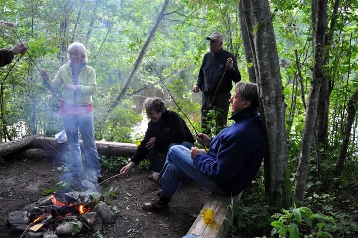 En grupp människor i jeans och fritidskläder sitter vid en lägereld omgiven av träd och skogsmiljö på Storängens Camping, Stugor & Outdoor.