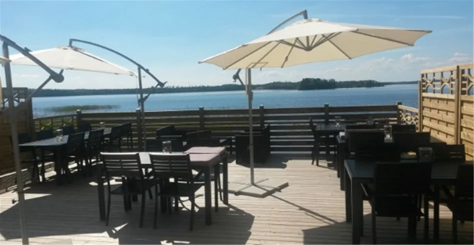 En campingplats med utomhusmöbler inklusive bord och stolar på en trädäck med utsikt över havet vid Vallsnäs Camping. Det finns skugga från parasoller och omgivningen har en avslappnad och inbjudande atmosfär.