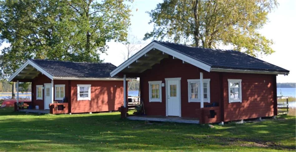 En pittoresk stuga omgiven av grönskande gräsmatta på Vallsnäs camping, med ett solbelyst tak och en skuggig gård, omgiven av natur och hus i närheten.