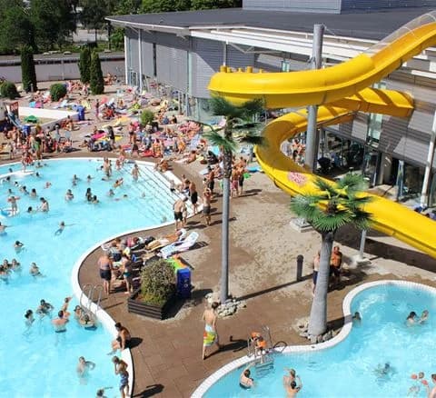 En livlig vattenpark med människor som njuter av semester och fritid, simmar i poolen och åker vattenrutschbanor. Flera personer bär badkläder och omgivningen utstrålar glädje och nöje.