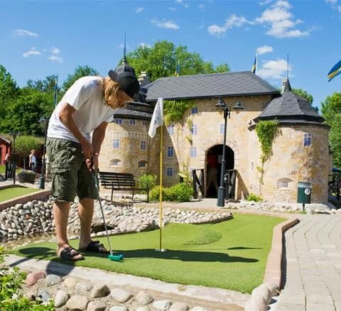 En grupp människor spelar minigolf på Gustavsvik Resort. Banan är omgiven av grönska och har färgglada hinder och utsmyckningar som skapar en rolig atmosfär.