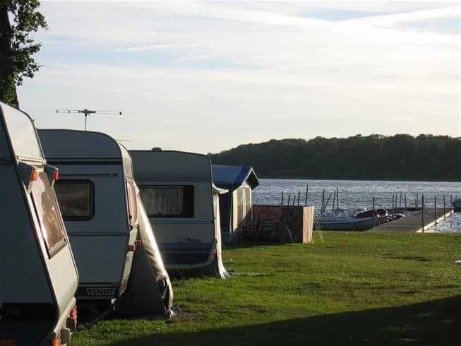 En idyllisk campingplats vid vattnet med husbilar och båtar vid en brygga, omgiven av naturskön miljö.