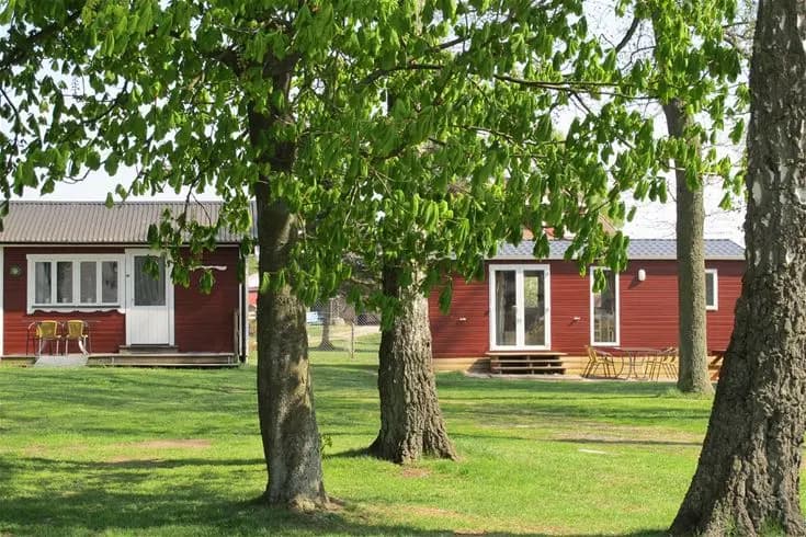 Bild av Jägersbo camping Swecamp: en grön gräsmatta omgiven av träd och buskar, med en mysig stuga i bakgrunden under en klar vårhimmel.