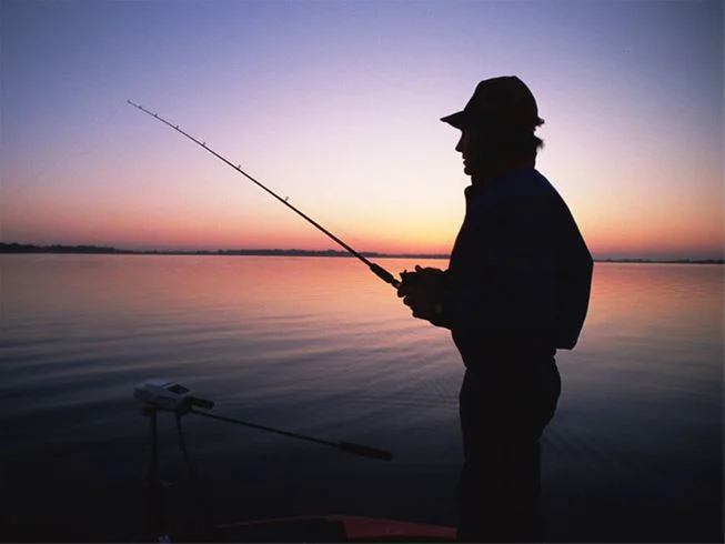 En silhuett av en fiskare står med ett fiskespö vid vattnet vid solnedgången på Degernäs camping, vilket skapar en stämningsfull och avslappnad atmosfär.